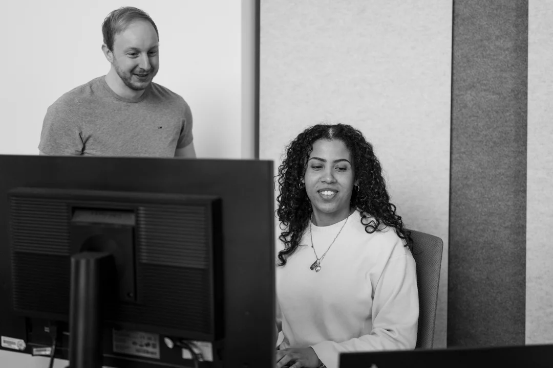 Ein Mann schaut in den Bildschirm einer Frau, die an einem Computer im Büro sitzt. Sie lächeln.