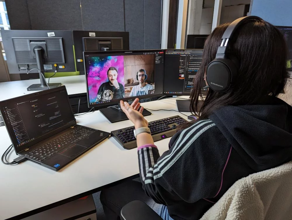 Lara sitzt mit Headset auf an ihrem Schreibtisch. Sie hat ein Videotelefonat mit zwei Kollegen.