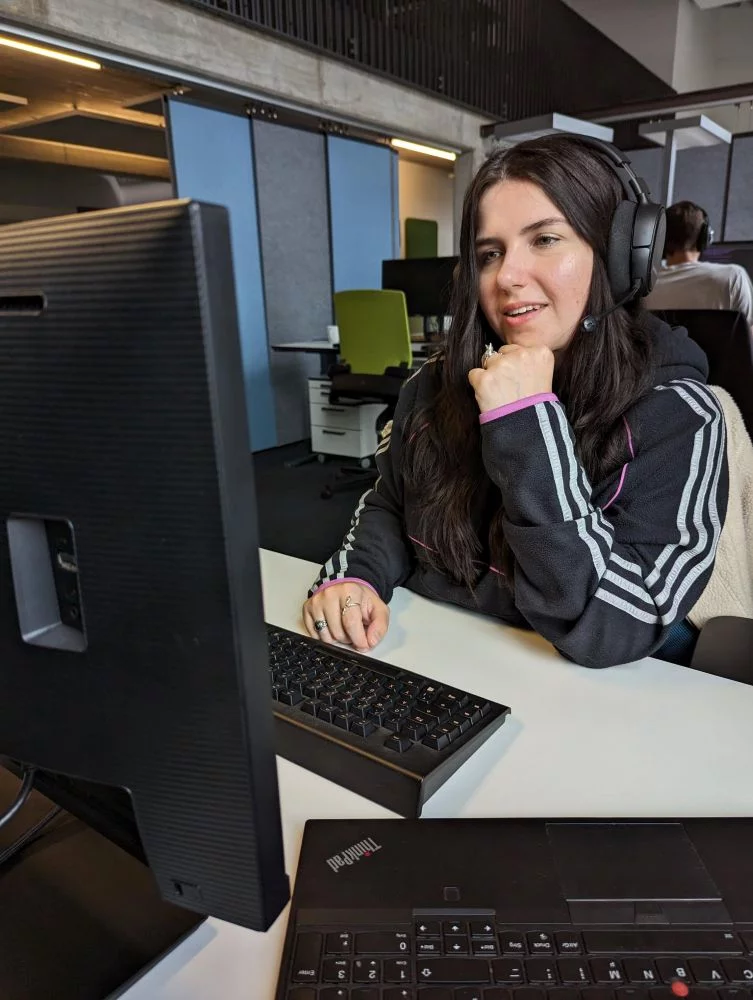 Lara sitzt an ihrem Schreibtisch mit Headset auf und spricht in die Kamera