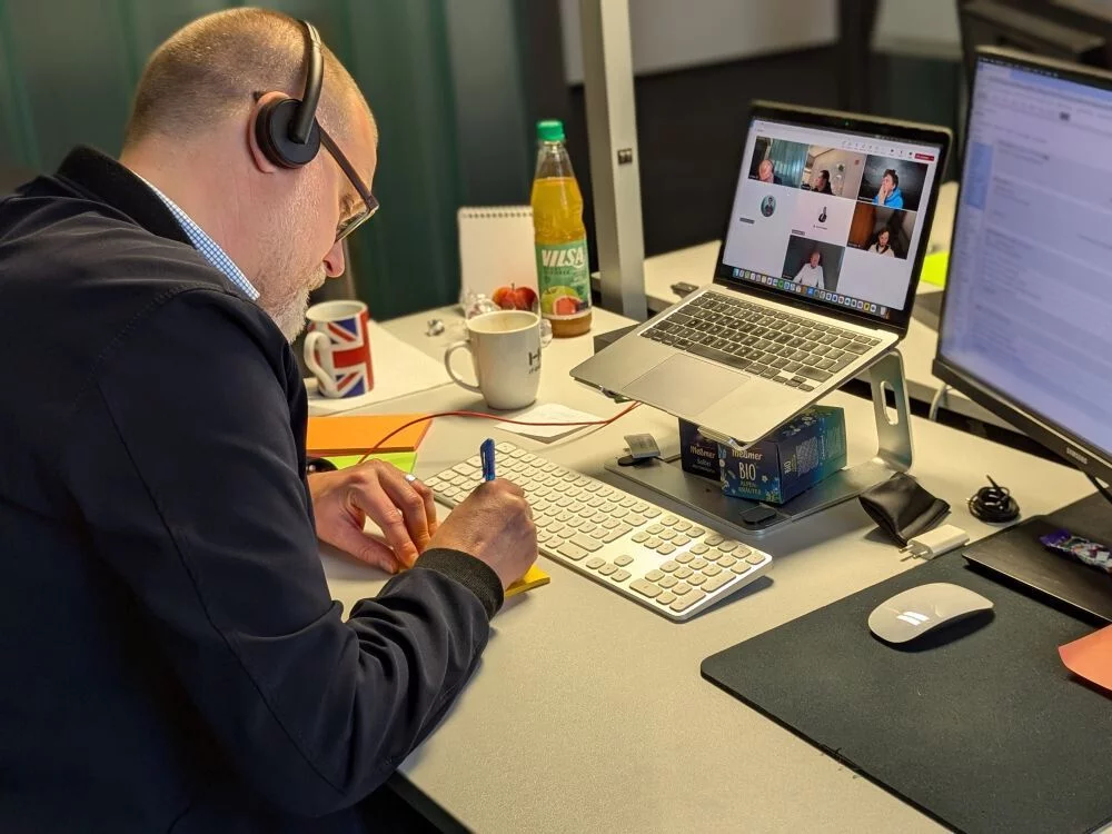 Tim sitzt am Schreibtisch, hat ein Headset auf und beschreibt Klebezettel