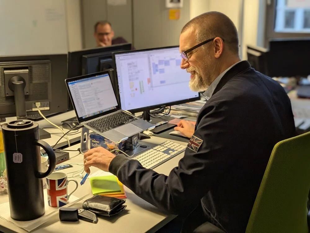 Tim sitzt an seinem Schreibtisch, checkt E-Mails und unterhält sich mit einem Kollegen.