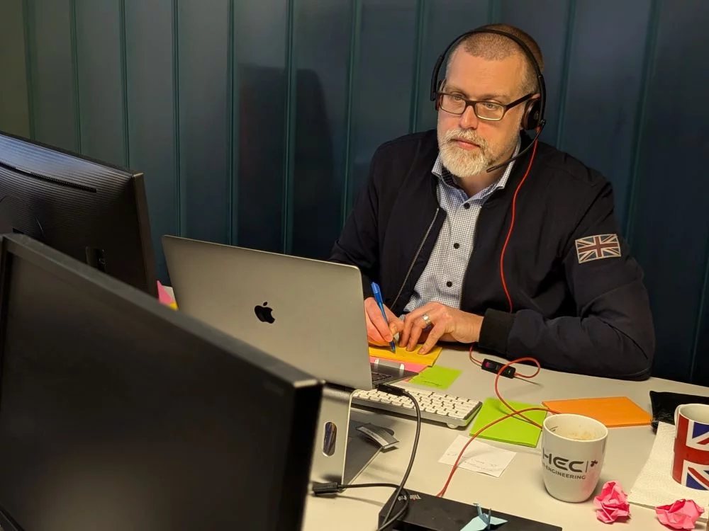 Tim sitzt am Schreibtisch, hat ein Headset auf und schreibt auf Klebezettel.