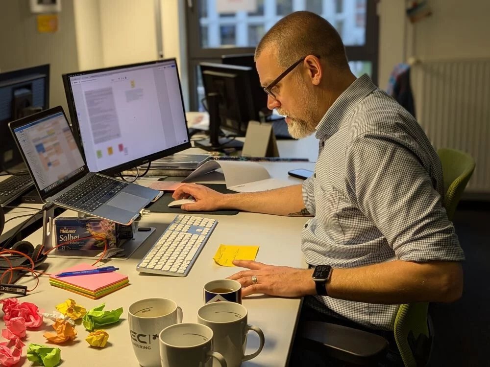 Tim sitzt am Schreibtisch, neben ihm liegen zerknüllte Klebezettel und stehen leere Kaffeebecher.