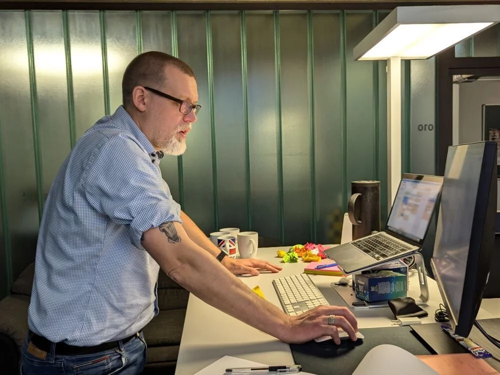 Tim steht vor seinem hochgefahrenen Schreibtisch, hat die Ärmel hochgekrempelt und klickt auf seine Mouse.