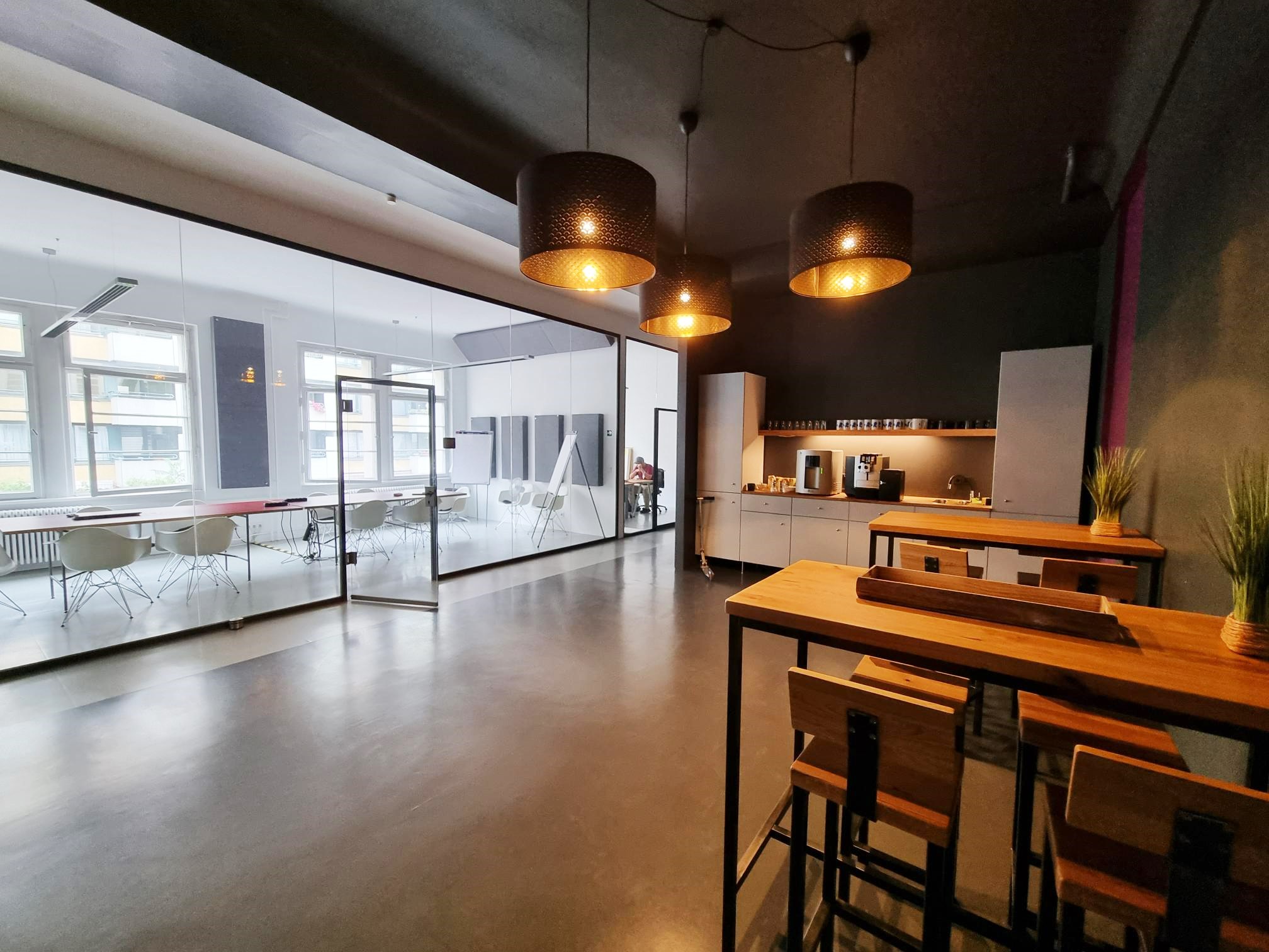 Auf der linken Seite ein großer gläserner Besprechungsraum, auf der rechten Seite davor eine Küche im Industriestil
