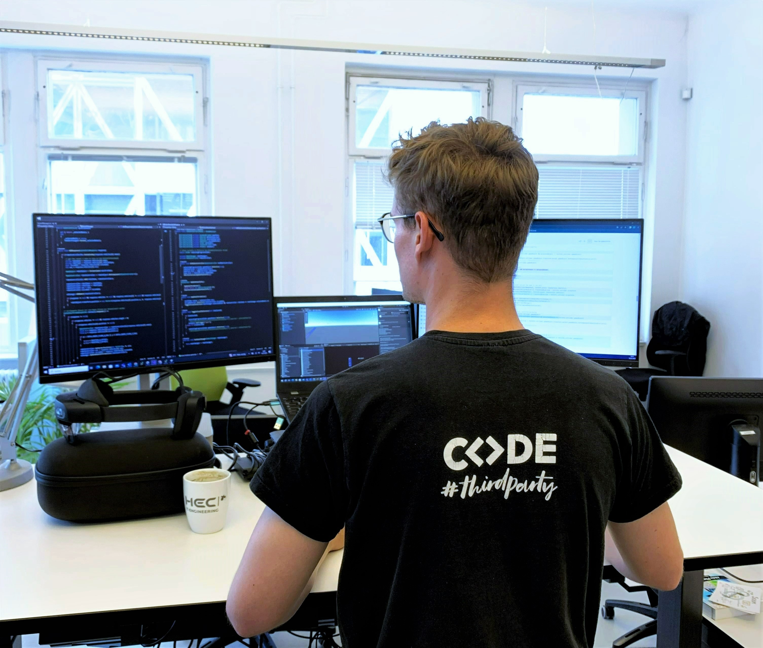 Werkstudent der HEC in Rückenansicht. Auf dem T-Shirt steht CODE. Er steht vor einem Schreibtisch. Werkstudent der HEC in Rückenansicht. Auf dem T-Shirt steht CODE. Er steht vor einem Schreibtisch.