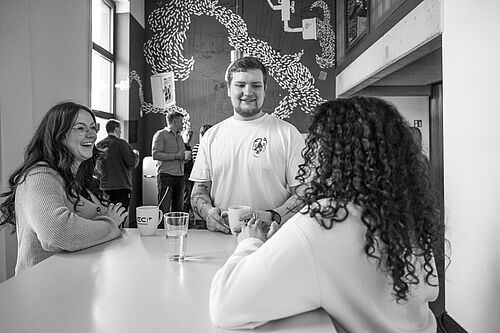 Mehrere junge Menschen stehen um einen Stehtisch in einem hohen, offenen Raum im Bürogebäude