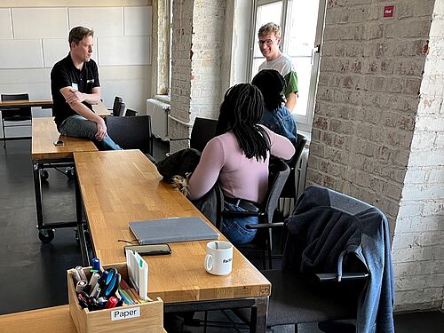 Seminarraum im Industriedesign. Hinten stehen zwei Männer, vorne sitzen zwei Frauen, die sich unterhalten.