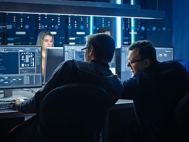 Zwei Männer sitzen vor Monitoren mit Code