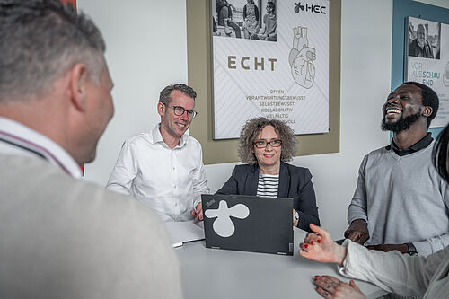 Mehrere  Menschen stehen um einen Stehtisch in einem Raum im Bürogebäude, sie lachen.
