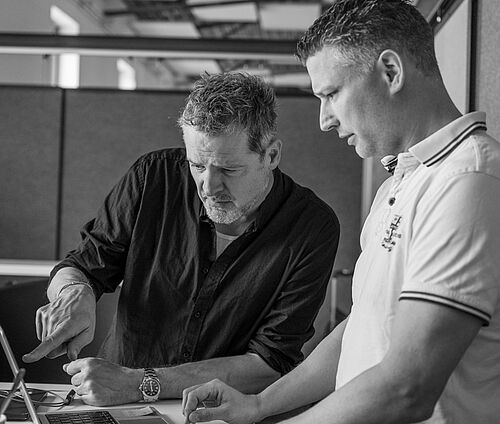 Ein Mann schaut in seinen Laptop Bildschirm. Er zeig einem anderen Mann etwas auf dem Bilschirm.