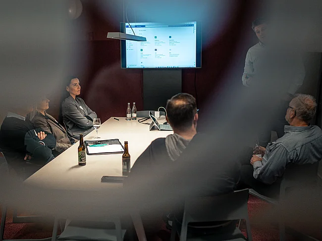 Durch eine Fensterscheibe sind Menschen an einem Schreibtisch zu sehen.