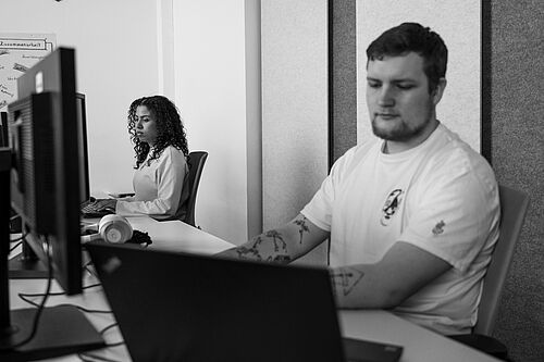 Ein Mann arbeitet an einem Computer Bildschirm im Büro der HEC. Im Hintergrund arbeitet auch eine Frau am PC.