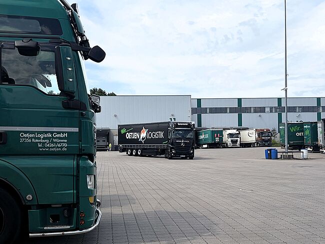 Im Vordergrund steht ein grüner LKW, im Hintergrund kommt ein schwarzer LKW über einen Hof gefahren.