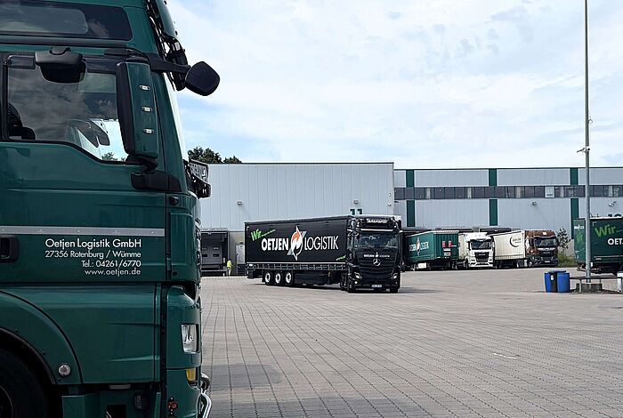 Im Vordergrund steht ein grüner LKW, im Hintergrund kommt ein schwarzer LKW über einen Hof gefahren.
