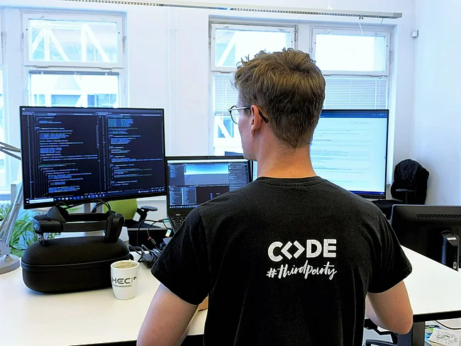 Werkstudent der HEC in Rückenansicht. Auf dem T-Shirt steht CODE. Er steht vor einem Schreibtisch.