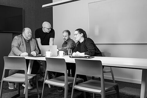 Drei Personen sitzen an einem Besprechungstisch, ein Mann steht und zeigtihnen etwas auf seinem Mobiltelefon