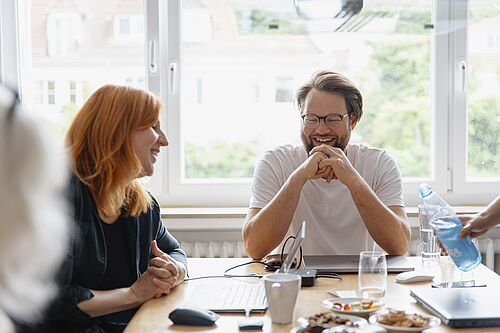 Eine Frau und ein Mann sitzen lachend an einem Arbeitstisch.