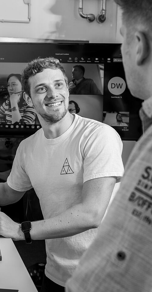 Zwei Männer in einer Besprechung, stehend, im Hintergrund ein großer Monitor mit Übertragung von Remote-Teilnehmenden in Kacheln