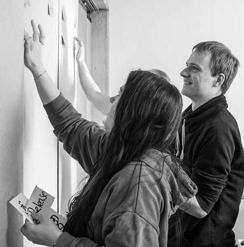 Mitarbeitende am Kanban Board Mehrere Personen stehen im Büro der HEC an einem Raumteiler, den sie mit beschriebenen Klebezetteln bestücken.