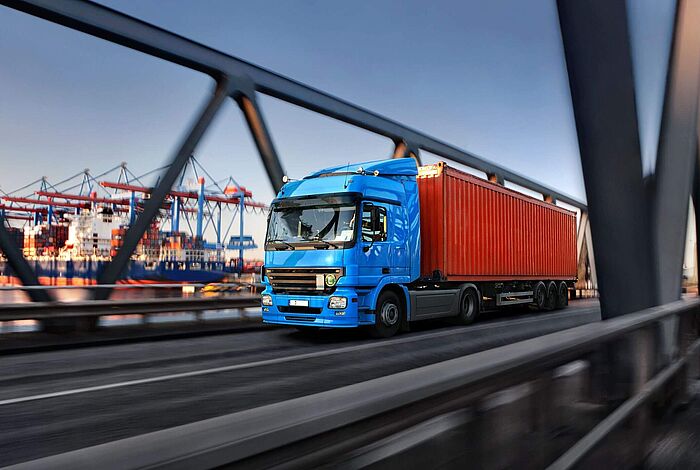 Ein LKW fährt über eine Brücke, im Hintergrund sind Hafenkräne zu sehen.