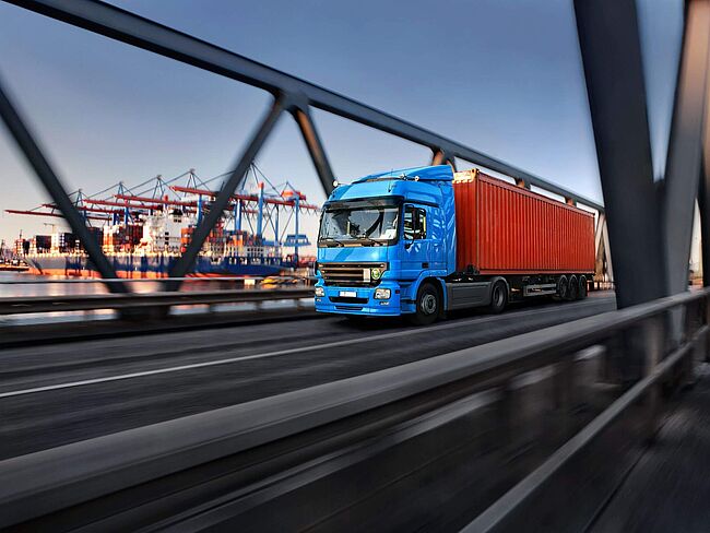 Ein LKW fährt über eine Brücke, im Hintergrund sind Hafenkräne zu sehen.