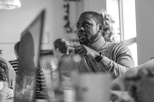 Ein Mann ist durch Meeting-Gegenstände hindurch an einem Stehtisch zu sehen, er redet und gestikuliert.