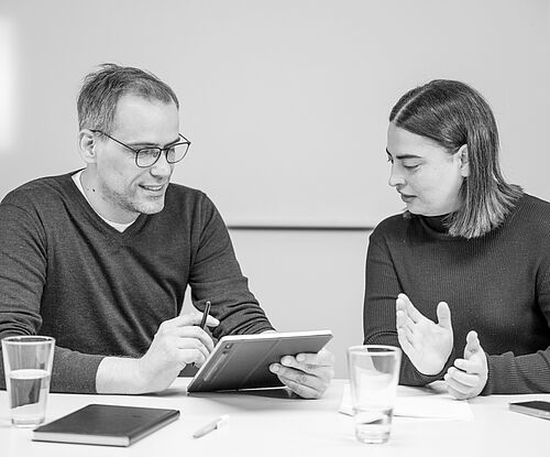 Ein Mann und eine Frau sitzen an einem Besprechungstisch und haben eine Diskussion vor einem Whiteboard
