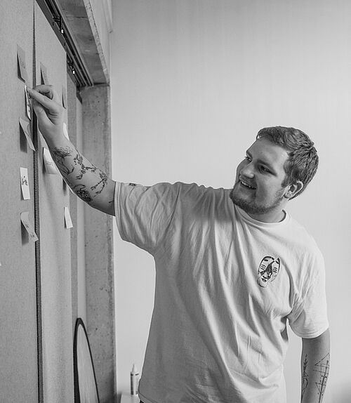 Zwei Personen stehen im Büro der HEC und arbeiten an einem Raumteiler, der mit beschriebenen Klebezetteln bestückt ist. 