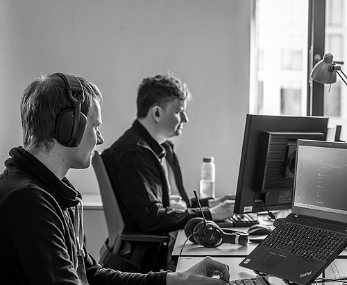 Zwei Männer schauen jeweils in ihren Computer Bildschirm im Büro der HEC. Sie tragen Headsets und sind fokussiert.