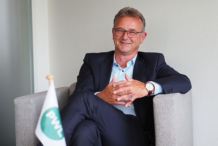 Ein Mann mit Anzug sitzt in einem Sessel. Vor ihm steht eine Flagge mit der Aufschrift PWL auf einem Tisch.