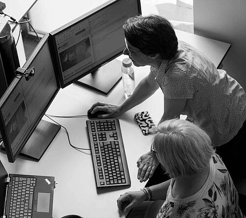 Zwei Frauen arbeiten gemeinsam vor zwei Monitoren an einem Laptop am Schreibtisch im Büro