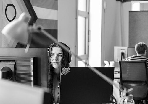Eine Frau arbeitet in einem Großraumbüro der HEC am Computer Bildschirm. Sie trägt ein Headset. An der Wand hängt eine Regenbogenfahne.