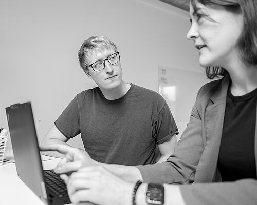 Eine Frau arbeitet am Laptop und redet mit einem Mann, der am nächsten Schreibtisch sitzt.