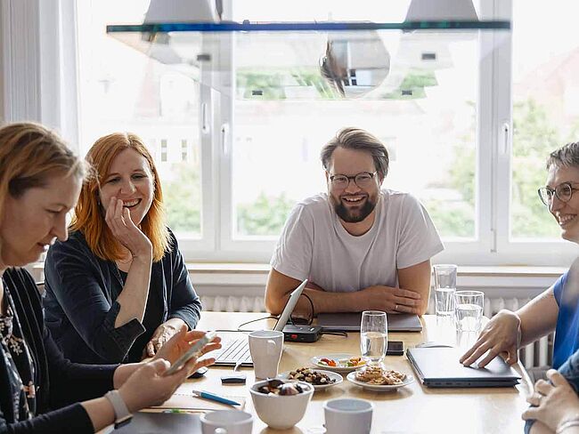 Frauen und ein Mann sitzen lachend an einem Arbeitstisch.	