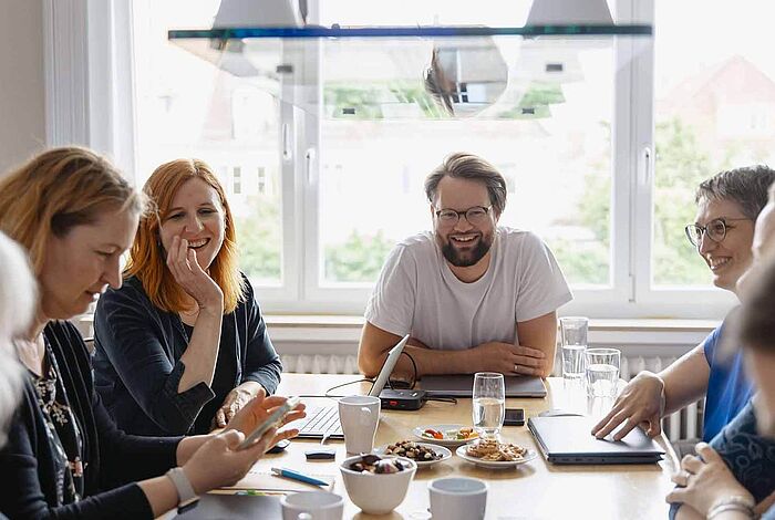 Frauen und ein Mann sitzen lachend an einem Arbeitstisch.	