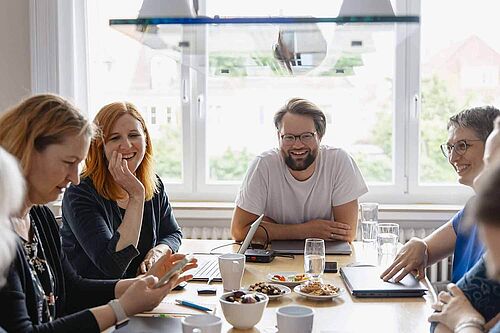 Frauen und ein Mann sitzen lachend an einem Arbeitstisch.	