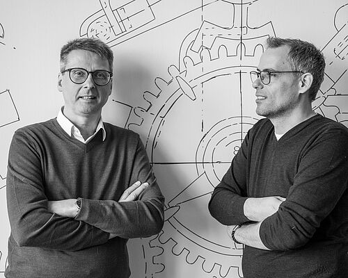 Zwei Männer lehnen jeweils mit verschränkten Armen an einer bemalten Wand.