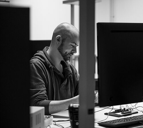 Ein Mann arbeitet zwischen anderen Schreibtischen auf denen Bildschirme stehen, an einem Gegenstand.