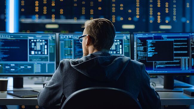 Ein Mann sitzt vor drei Computermonitoren mit Code
