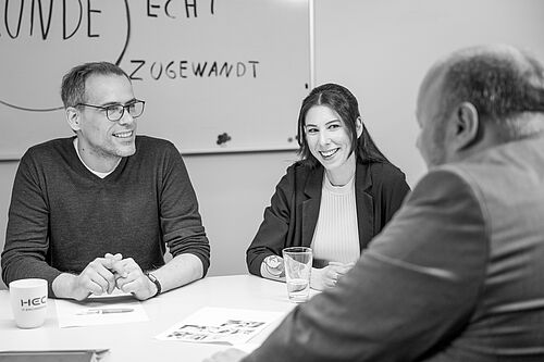 Drei Personen sitzen an einem Besprechungstisch, reden und lächeln.