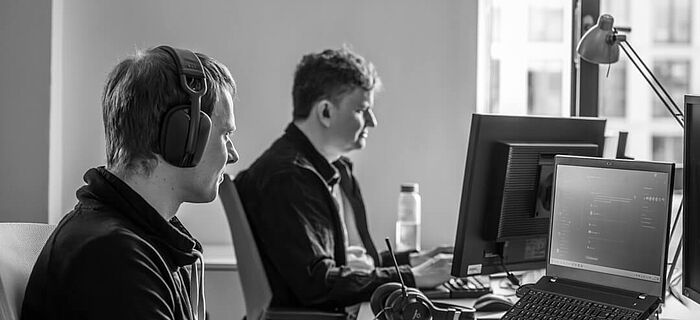 Zwei Männer schauen jeweils in ihren Computer Bildschirm im Büro der HEC. Sie tragen Headsets und sind fokussiert.