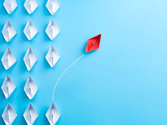 Weiße Papierschiffe sind gleichmäßig auf einem blauen Grund angeordnet. Ein rotes Papierschiff schert aus der Reihe aus.