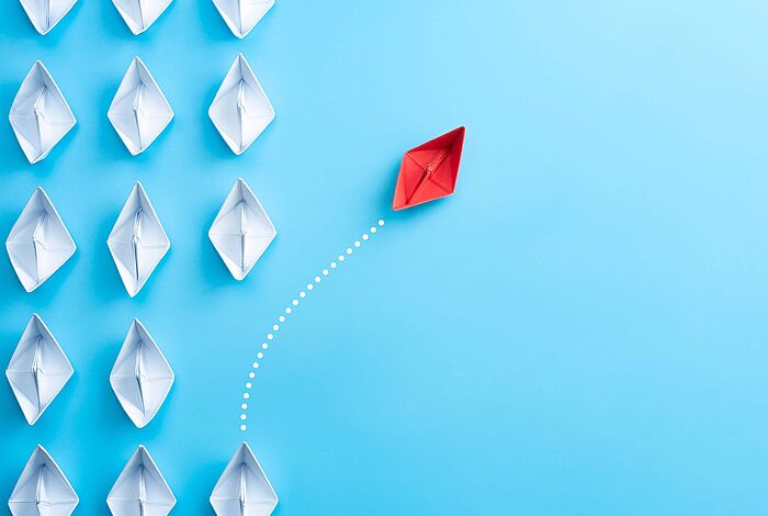 Weiße Papierschiffe sind gleichmäßig auf einem blauen Grund angeordnet. Ein rotes Papierschiff schert aus der Reihe aus.