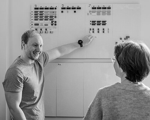 Ein Mann steht an einem Planning Board an der Bürowand und zeigt auf eine Stelle. Eine Frau sieht ihm zu. 