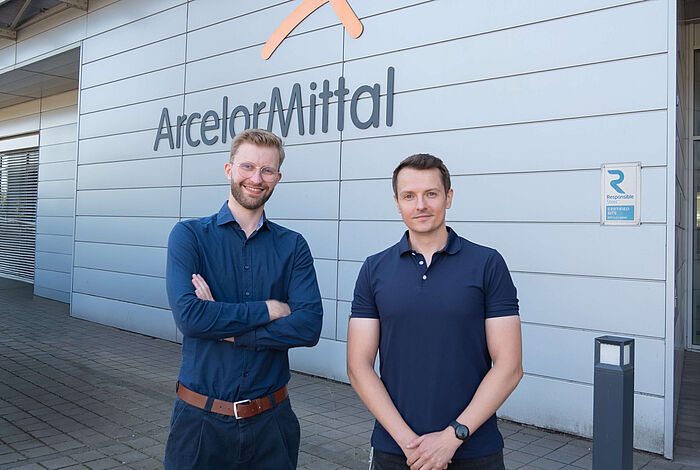 Zwei Männer stehen vor einer Wand mit dem Logo von ArcelorMittal Bremen und lächeln