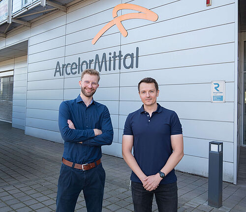 Zwei Männer stehen vor einer Wand mit dem Logo von ArcelorMittal Bremen und lächeln