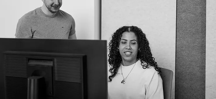 Ein Mann schaut in den Bildschirm einer Frau, die an einem Computer im Büro sitzt. Sie lächeln.