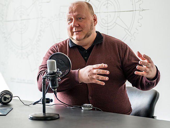 Ein Mann sitzt vor einem Mikrofon am Tisch, redet und gestikuliert