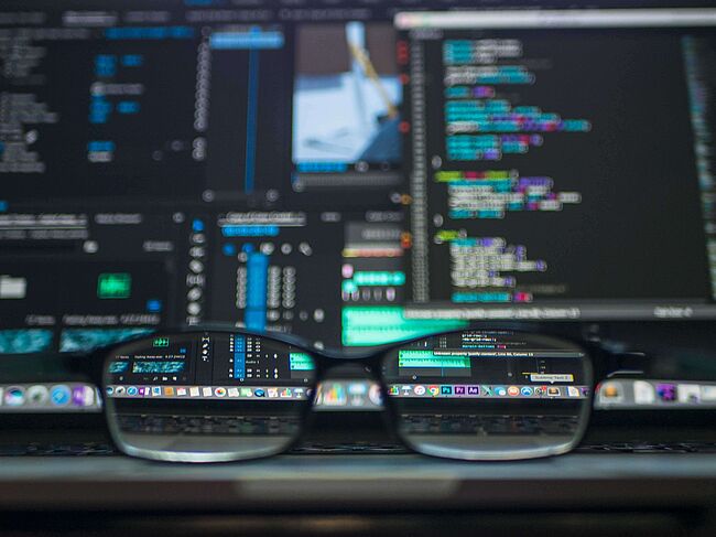 Im Hintergrund ist ein Bildschirm voller Daten und Algorithmen. Im Vordergrund erkennt man eine Brille. 
